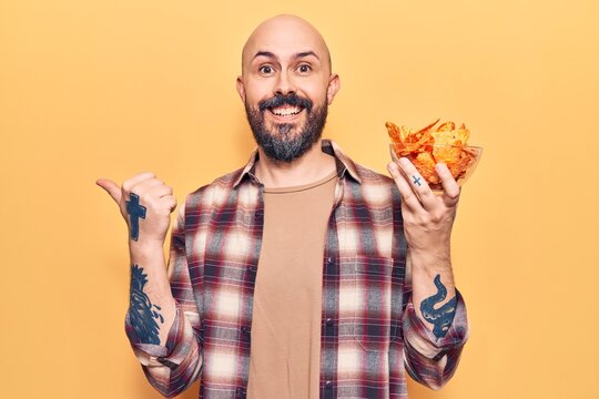 Young handsome man holding nachos potato chips pointing thumb up to the side smiling happy with open mouth