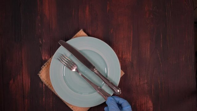 Women's gloved hands place a metal knife and fork on an empty plate on a brown wooden table.