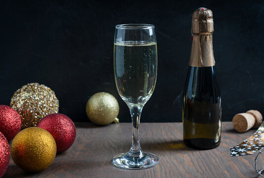 Wineglass full of dry white Prosecco sparkling champagne with golden and red christmas or new year decoration balls and bottle on dark brown wooden table against black background. Horizontal image