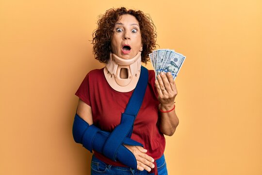 Beautiful middle age mature woman wearing cervical collar and sling holding insurance dollars afraid and shocked with surprise and amazed expression, fear and excited face.