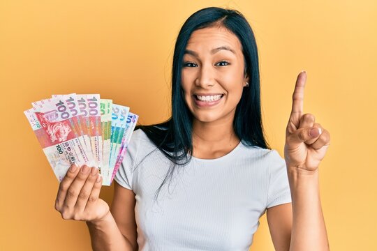 Beautiful hispanic woman holding hong kong dollars banknotes smiling with an idea or question pointing finger with happy face, number one