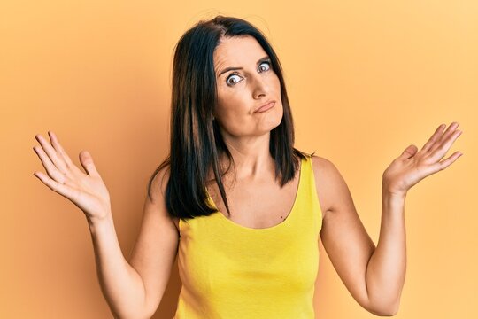 Middle age brunette woman wearing casual clothes over yellow background clueless and confused with open arms, no idea and doubtful face.