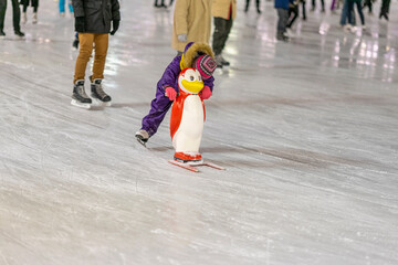 Ice rink. Little child holds a penguin for support and learn to skate. Happy childhood and Winter activities
