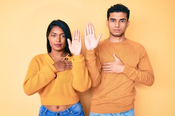 Beautiful latin young couple wearing casual clothes together swearing with hand on chest and open...
