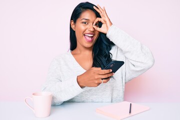 Beautiful latin young woman with long hair using smartphone sitting on the table smiling happy doing ok sign with hand on eye looking through fingers