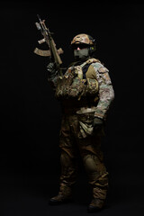 Full length photo of army soldier standing in full military uniform, wearing bulletproof vest, helmet, glasses and mask, holding up machine gun and looking at camera, isolated on black background