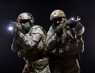 Two soldiers in military gear and bulletproof vests cover each other and raise their submachine...