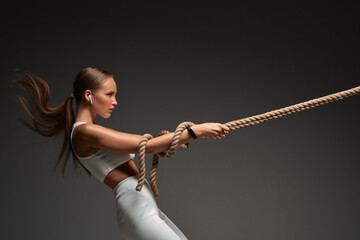 Side view of sporty attractive woman in white sportswear pulling rope isolated on gray background.