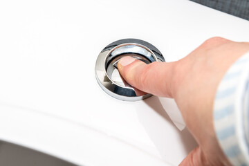 Close up of finger is pushing on a shiny metal flush button on white ceramic toilet bowl for cleaning. Save water concept.