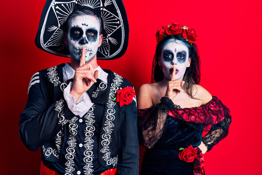 Young couple wearing mexican day of the dead costume over red asking to be quiet with finger on lips. silence and secret concept.
