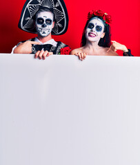 Young couple wearing mexican day of the dead costume holding blank empty banner pointing finger to one self smiling happy and proud