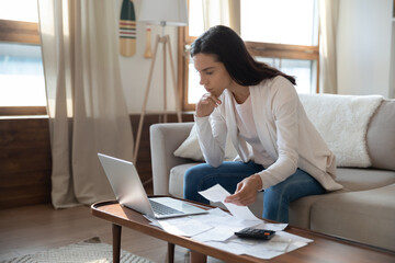 How to save more money. Anxious young woman looking at laptop screen holding invoice receipt in...