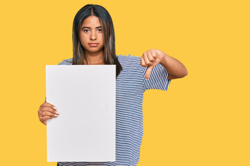 Young latin girl holding blank empty banner with angry face, negative sign showing dislike with...