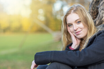 Smiling young woman is posing in the autumn park supporting her head with  hand looking at the...