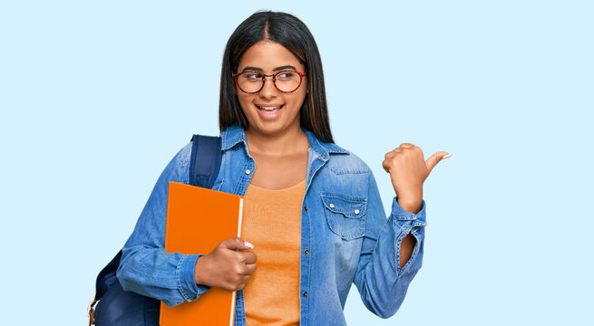 Young latin girl wearing student backpack and holding books smiling with happy face looking and pointing to the side with thumb up.