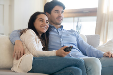 Relaxing by screen. Happy young spouses or couple in love cuddling on soft couch at home resting...