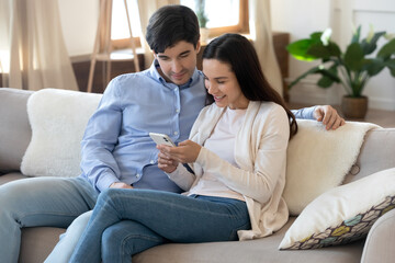 Online together. Happy loving young family couple sitting on couch browsing internet or social networks using cellphone, making video call, chatting with friends, watching cute photos on gadget screen