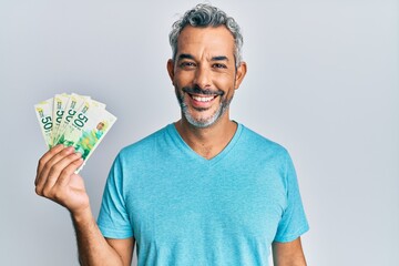 Middle age grey-haired man holding israel shekels banknotes looking positive and happy standing and smiling with a confident smile showing teeth