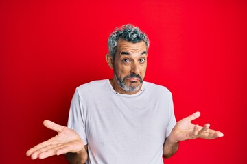 Middle age grey-haired man wearing casual white tshirt clueless and confused expression with arms and hands raised. doubt concept.