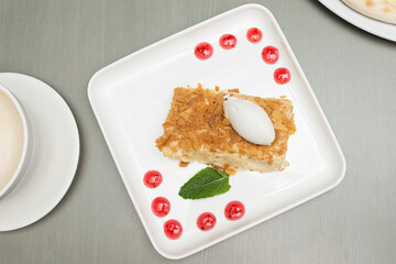 Slice of Napoleon cake and cocoa on gray background. Slice of ice cream on the cake and cappuccino on the table. A piece of cake on a square plate decorated with drops of red jam and a leaf of mint.