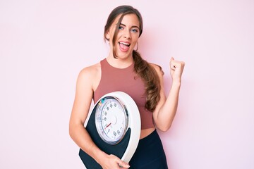Beautiful young caucasian woman holding weight machine to balance weight loss screaming proud,...