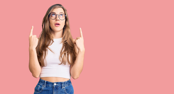Beautiful caucasian young woman wearing casual clothes and glasses amazed and surprised looking up and pointing with fingers and raised arms.