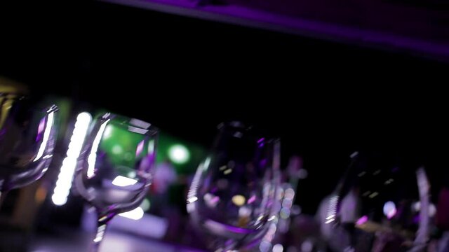 The concert stage lights reflect in empty clean glasses on an open counter. Multicolored lights of light in low light. Wine glasses at the party. Free camera movement. 