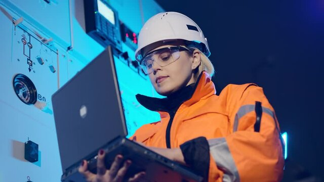Woman in helmet and orange uniform with laptop checks new equipment data at wind power plant substation close low angle shot