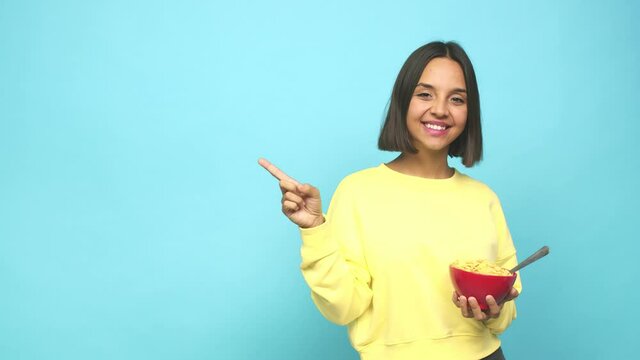 Young latin woman eating a cereals bowl smiling and pointing aside, showing something at blank space