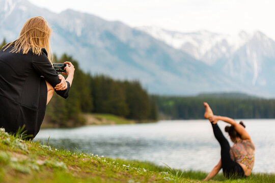 Young woman taking photo of a young fitness girl doing yoga pose by the lake. Women relaxing and having fun. Concept about lifestyle, people, sport and nature. 