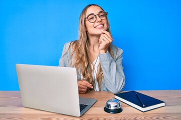 Young beautiful blonde woman sitting at hotel reception smiling looking confident at the camera with crossed arms and hand on chin. thinking positive.