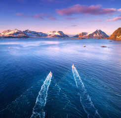 Aerial view of two fishing boats, rocks in the blue sea, snowy mountains and colorful sky with...