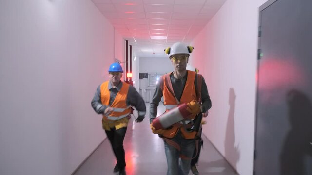 Worker with fire extinguisher and colleagues in orange vests run along hallway at warning signal flashes due accident at plant