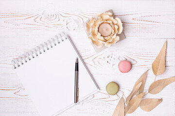 Top view on blank album and metal pen, beige leaves, macaroons and flower-shaped candlestick on a white wooden table