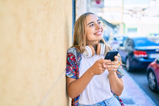 Young beautiful blonde caucasian woman smiling happy outdoors on a sunny day wearing headphones and using smartphone
