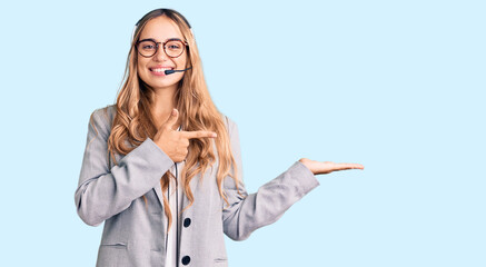 Young beautiful blonde woman wearing call center agent headset amazed and smiling to the camera while presenting with hand and pointing with finger.