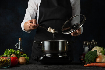 Chef in black apron testing hot soup at the professional restaurant kitchen on dark blue background. Backstage of preparing dish for dinner. Coking process concept. Cookbook.