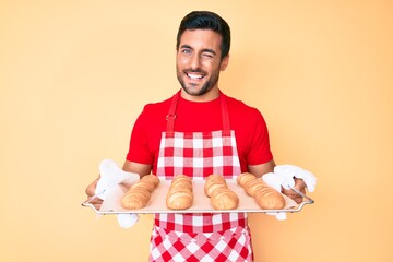Young hispanic man wearing baker apron holding homemade bread winking looking at the camera with...