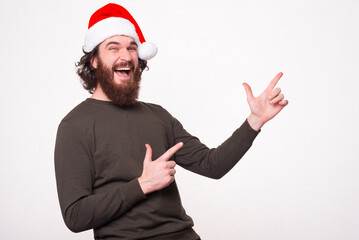 Joyful amazed bearded hipster man wearing santa claus hat and pointing at copyspace