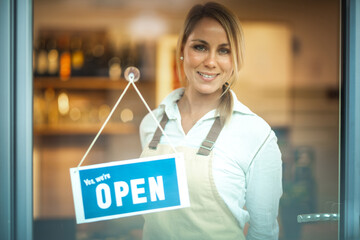 Portrait of a smiling Caucasian woman with beige apron, standing behind glass door, with sign ''we are open'' 