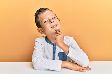 Adorable caucasian kid wearing casual clothes sitting on the table looking confident at the camera with smile with crossed arms and hand raised on chin. thinking positive.