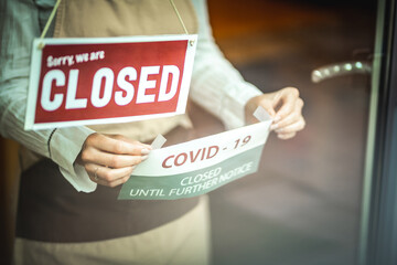 Female silhouette with a beige apron, standing behind the glass door with the sign ''closed'' and putting the covid19 notice