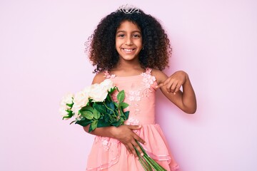 African american child with curly hair wearing princess crown holding flowers smiling happy pointing with hand and finger © Krakenimages.com