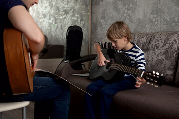 Young male musician teaches little boy student how to play the acoustic guitar and play by notes