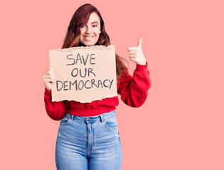 Young beautiful woman holding save our democracy protest banner smiling happy and positive, thumb up doing excellent and approval sign