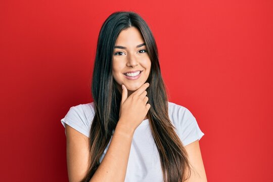 Young brunette woman wearing casual white tshirt over red background looking confident at the camera smiling with crossed arms and hand raised on chin. thinking positive.