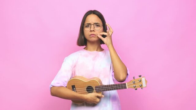 Young latin woman playing ukelele with fingers on lips keeping a secret