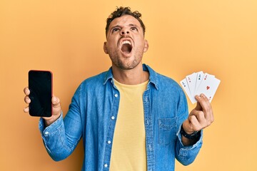 Handsome man with beard holding poker cards and smartphone showing blank screen winking looking at...