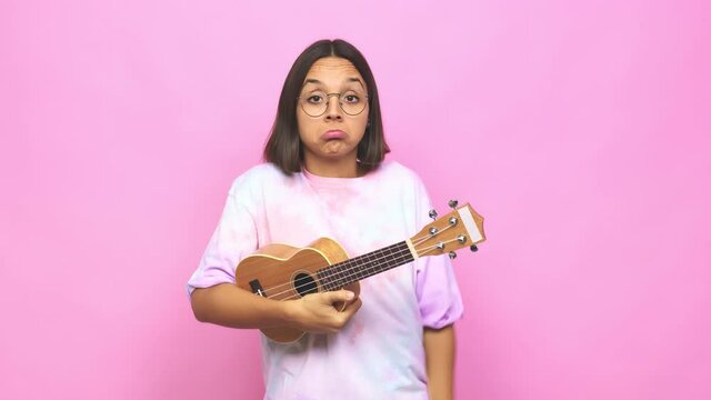 Young latin woman playing ukelele shrugs shoulders and open eyes confused
