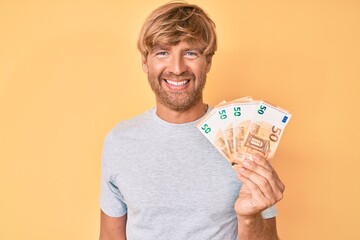 Young blond man holding euro banknotes looking positive and happy standing and smiling with a...